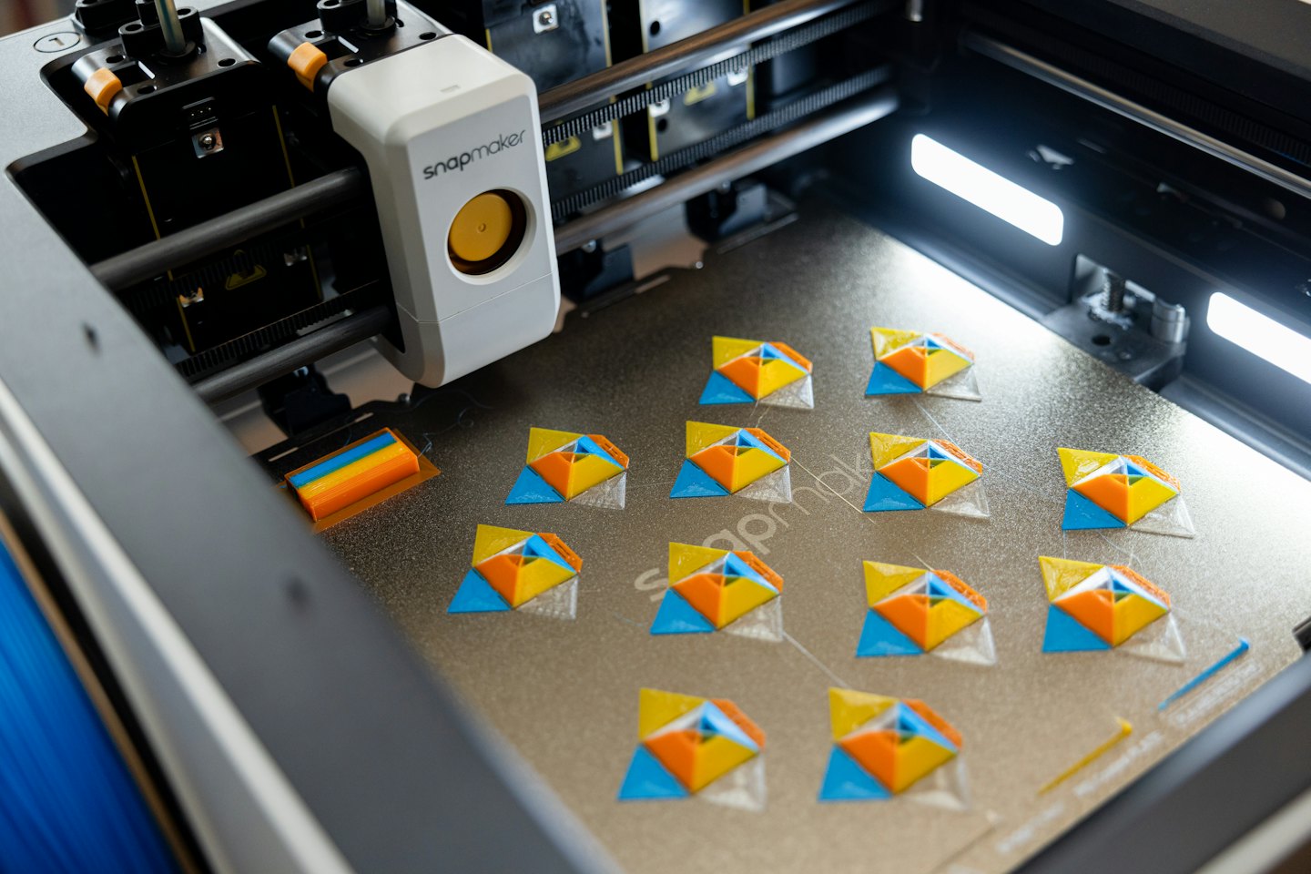 Drukarka 3D tworzy małe niebieskie, pomarańczowe i żółte obiekty geometryczne.