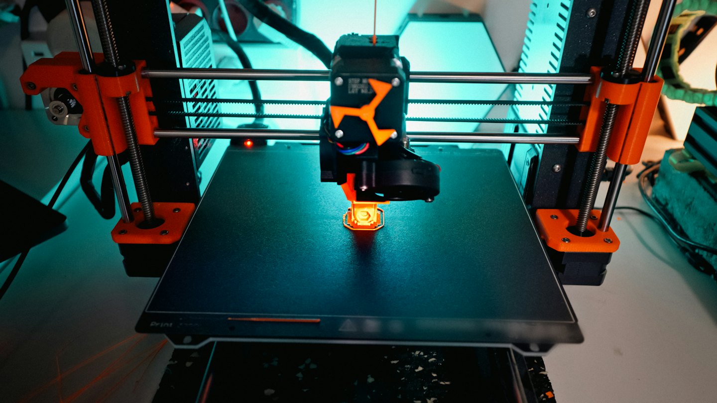 Drukarka 3D wytłacza świecący pomarańczowy plastik na platformę roboczą.